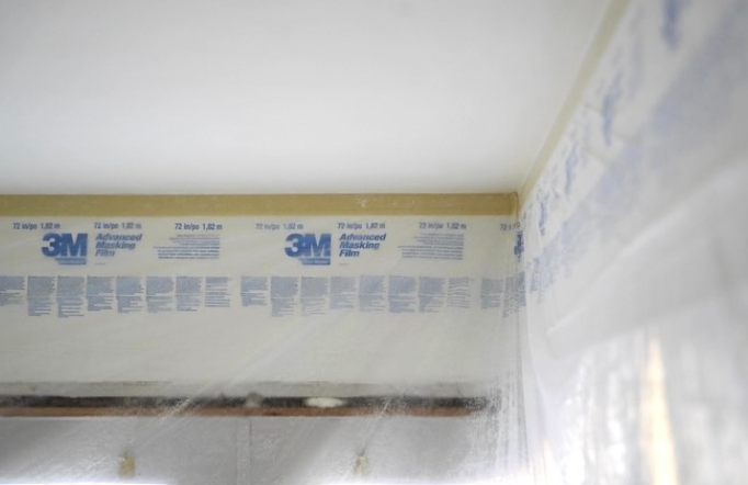 The corner of a room is lined with masking film, plastic sheeting, and blue printed tape along the wall and ceiling.