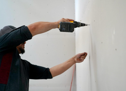 A person is using a cordless drill on a white wall while holding a small fastener against the drywall.