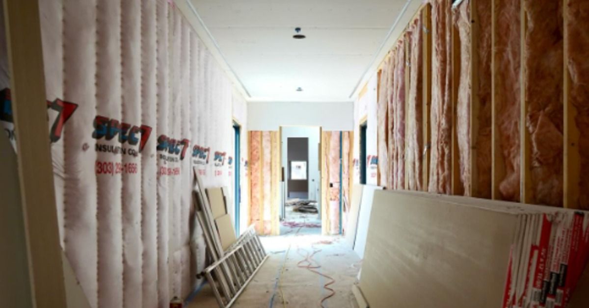 A hallway under construction with exposed insulation, drywall sheets, a ladder, tools, and wires on the floor.