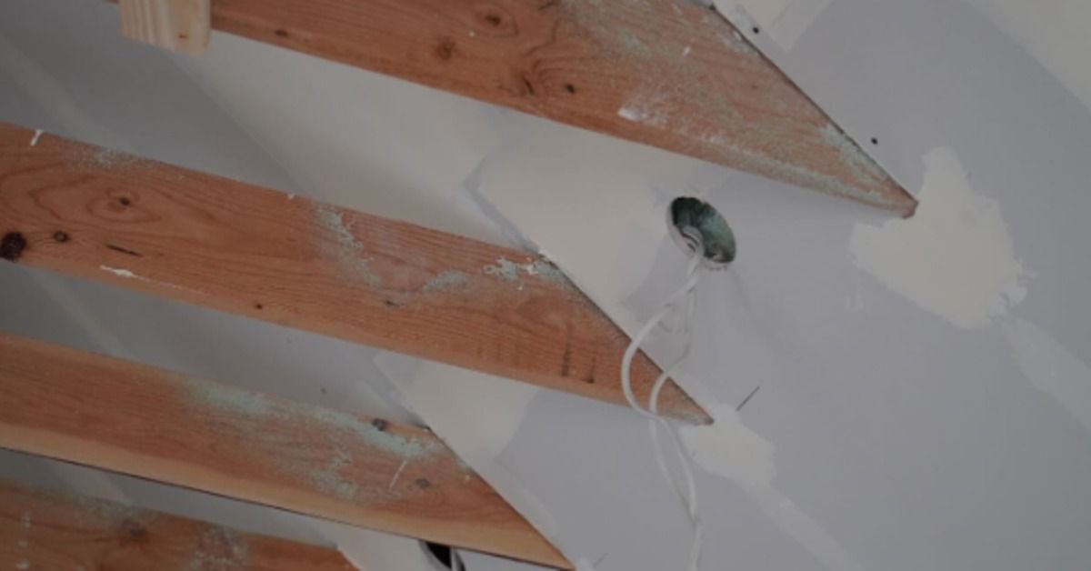 A close-up of exposed wooden beams on a gray ceiling. Loose wires are hanging from a circular light opening.