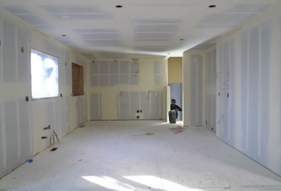 A partially finished room with drywall panels, ceiling cutouts, exposed wires, and a worker standing