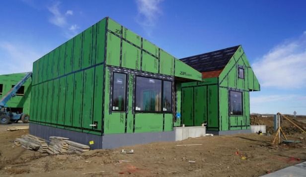 A house under construction with green exterior sheathing, black-framed windows, an open entryway, and exposed roof framing.