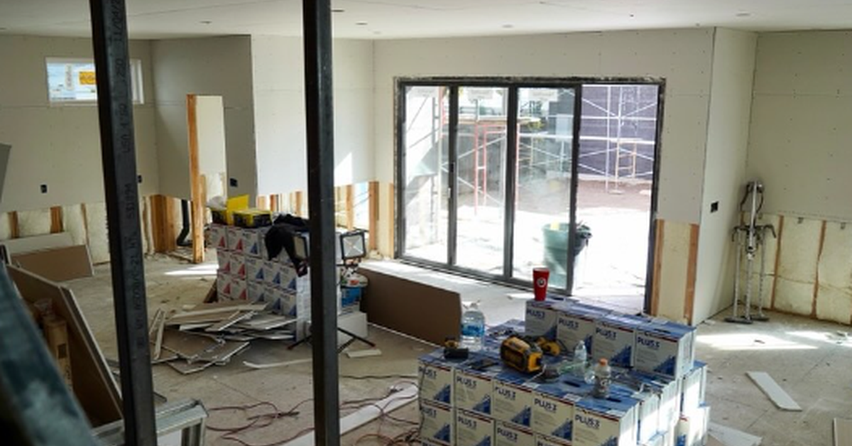 A worker uses a power tool beside the frame of the ceiling. Drywall, exposed pipes, and unfinished w