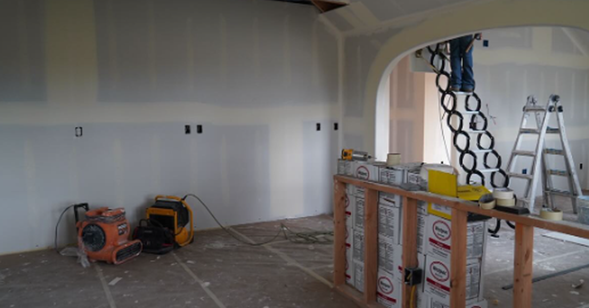 An unfinished interior with drywall panels, support columns, boxed materials, tools, and debris across the floor.