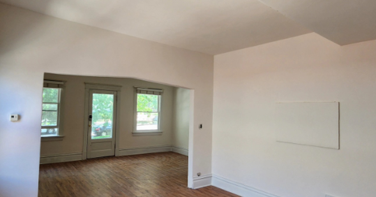 An empty room with wood plank flooring and white walls leading into a small space with two windows a