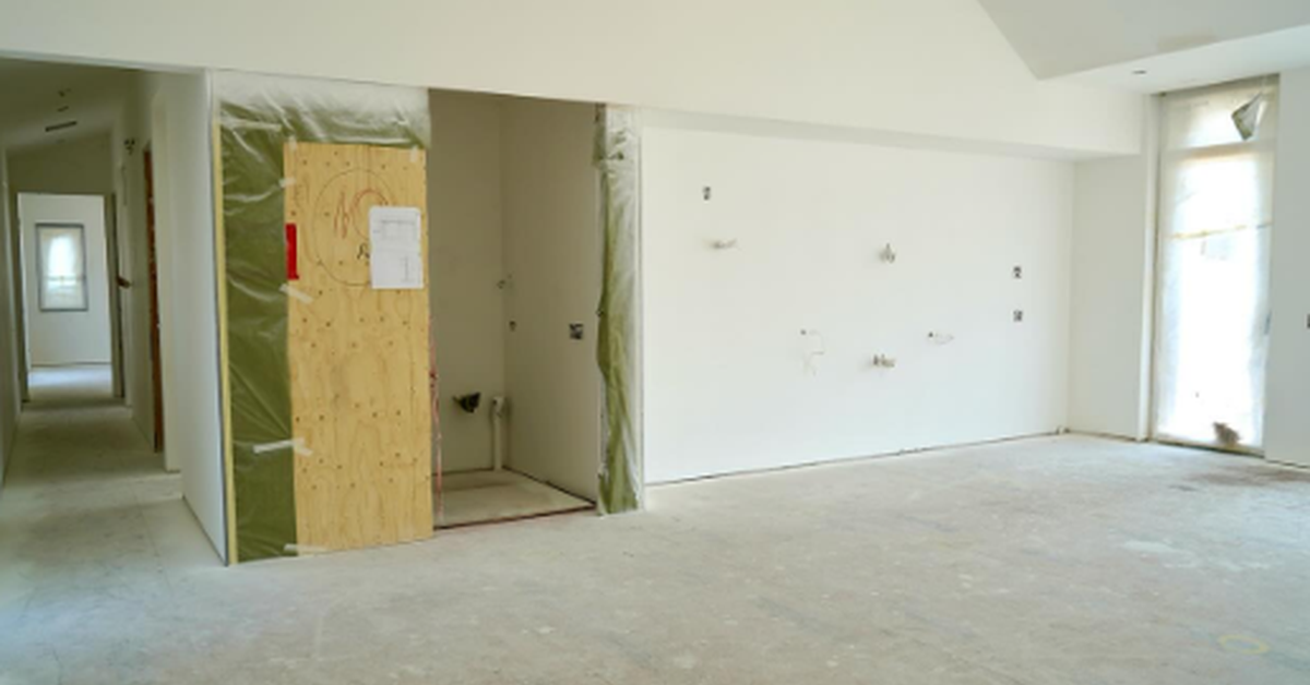 An unfinished interior room with bare concrete flooring, white walls, and a plywood-covered opening beside a hallway.