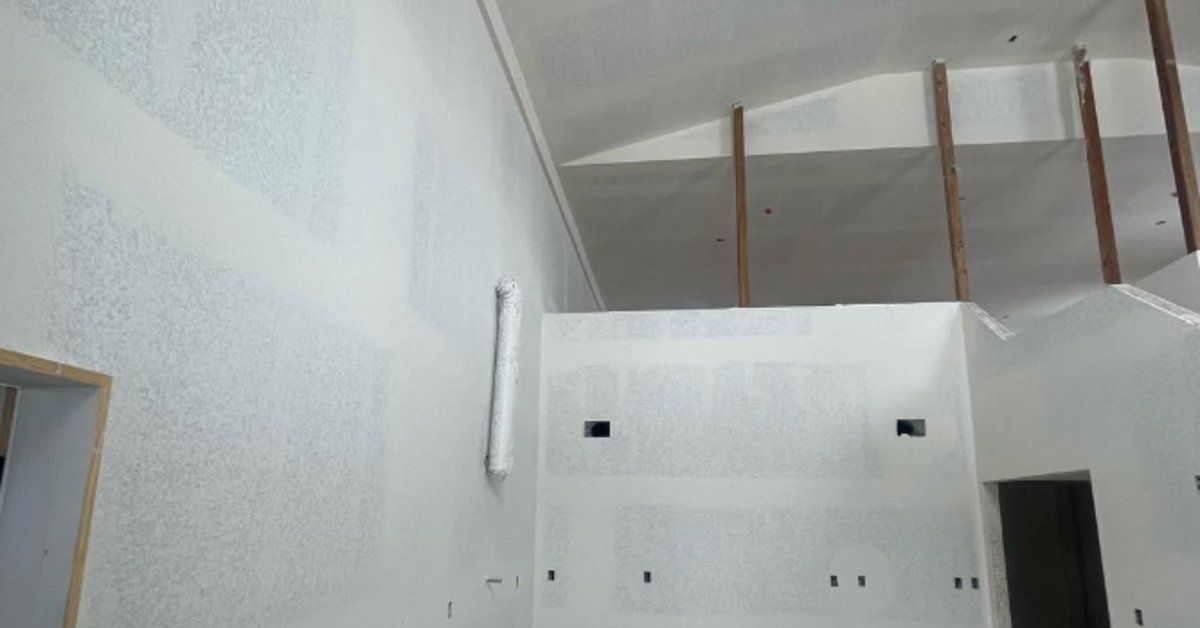 A high-ceilinged interior room with newly installed drywall, exposed beams, and unfinished construct