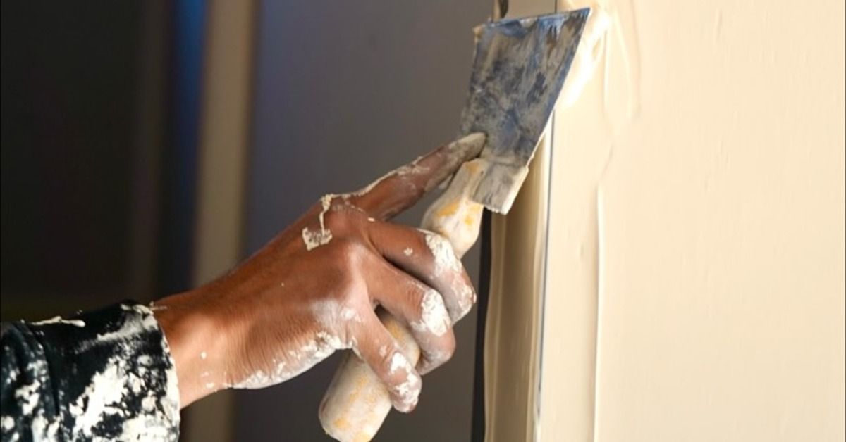 A close-up of a mud-covered hand holding a metal putty knife against a smooth beige wall indoors bes