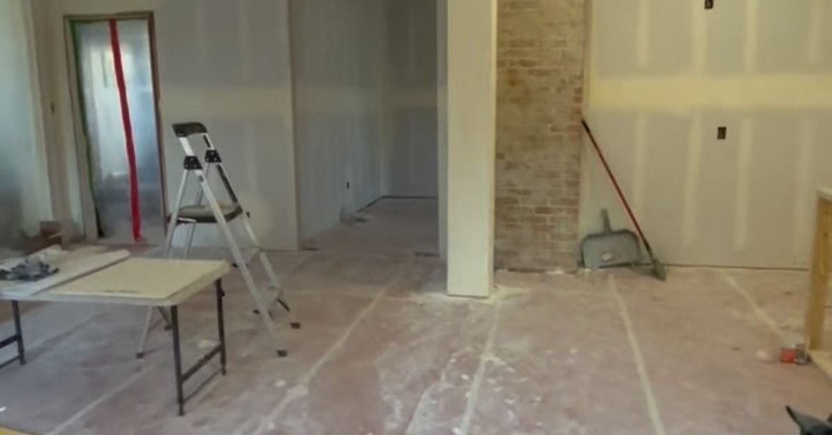 An unfinished room with a ladder, table, broom, and dusty floor in front of partially finished drywall.