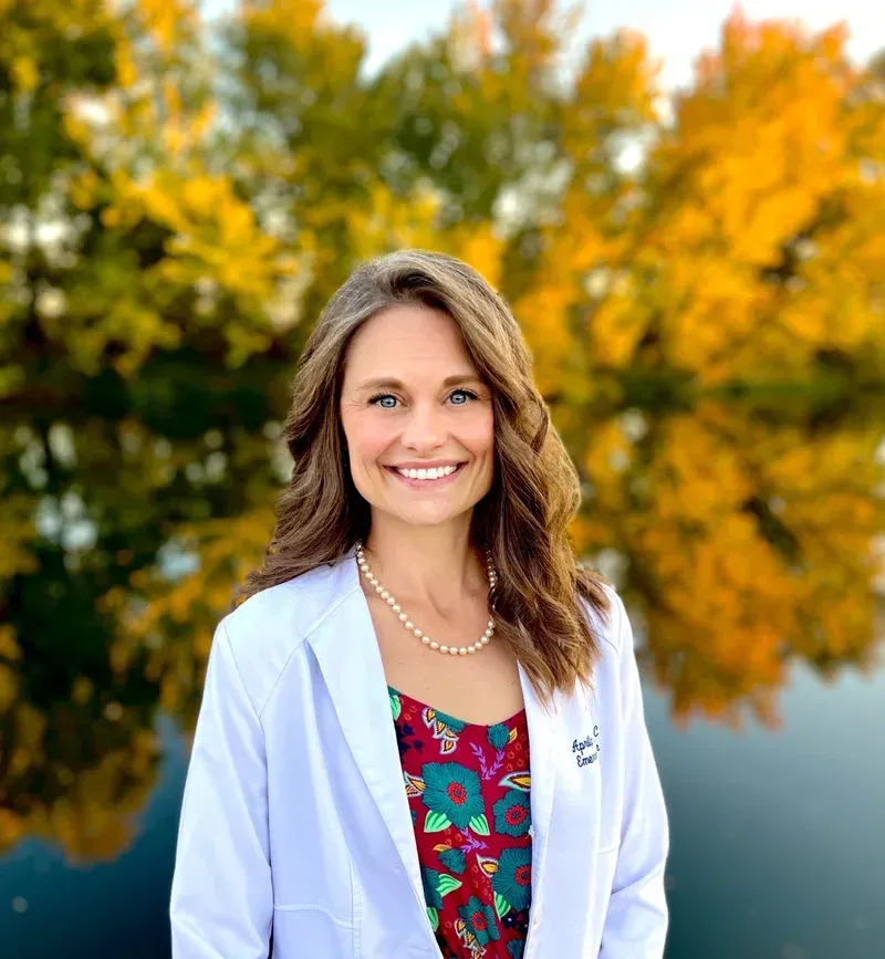A woman in a white coat is smiling in front of a lake.