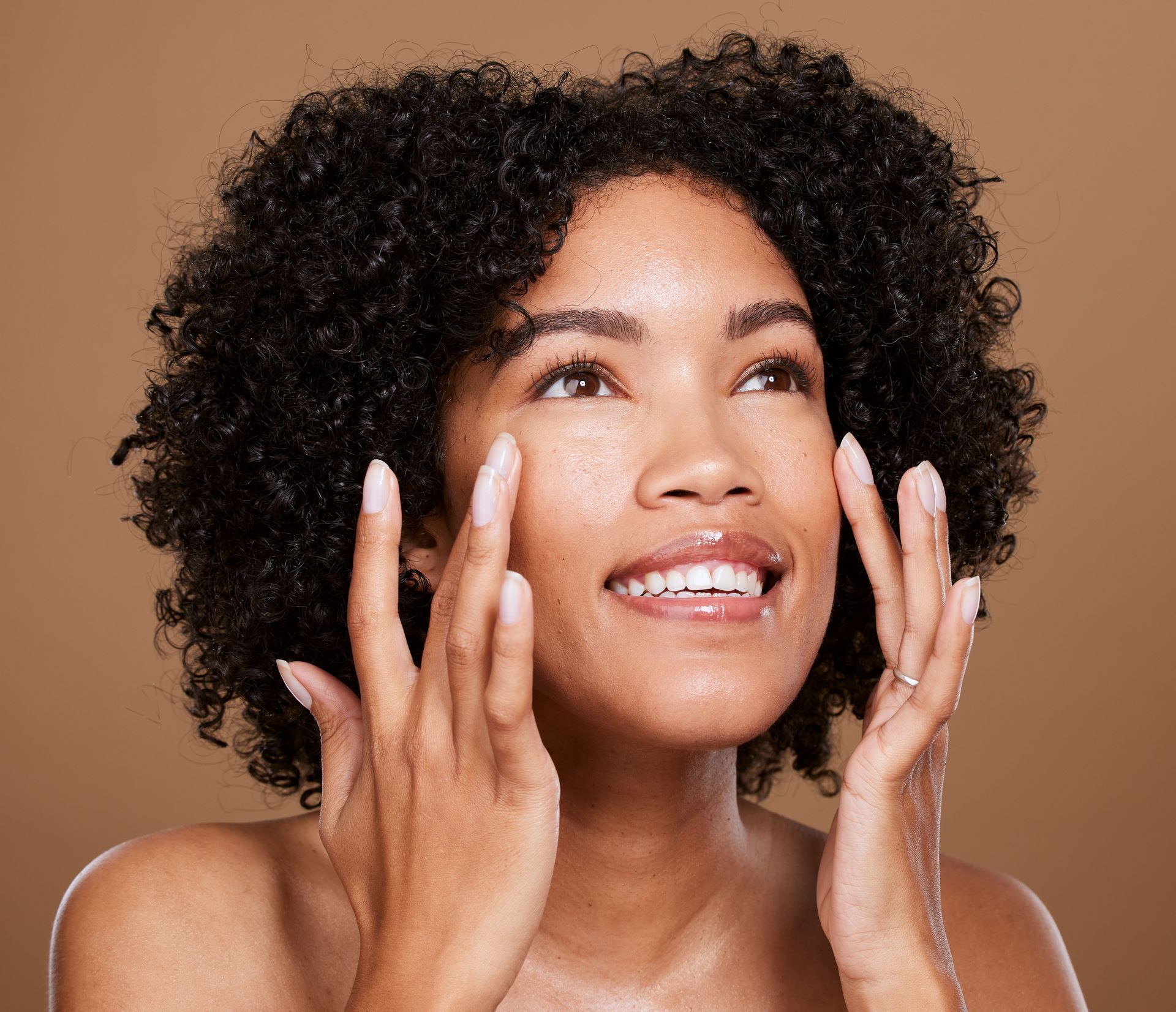 Smiling person with curly hair touching their face against a beige background