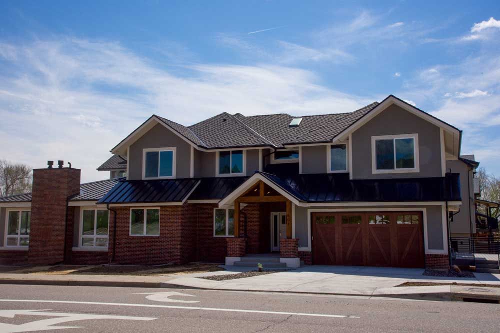 Two-story house with gray siding, brick, black roof, and wooden garage door against a blue sky.