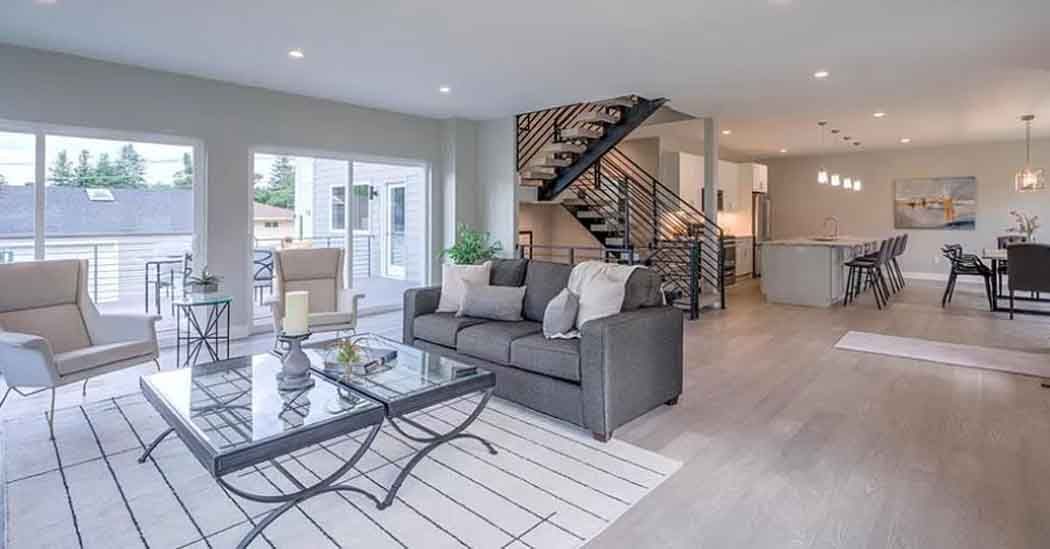 Modern living room with gray sofa, staircase, dining area, and large windows.