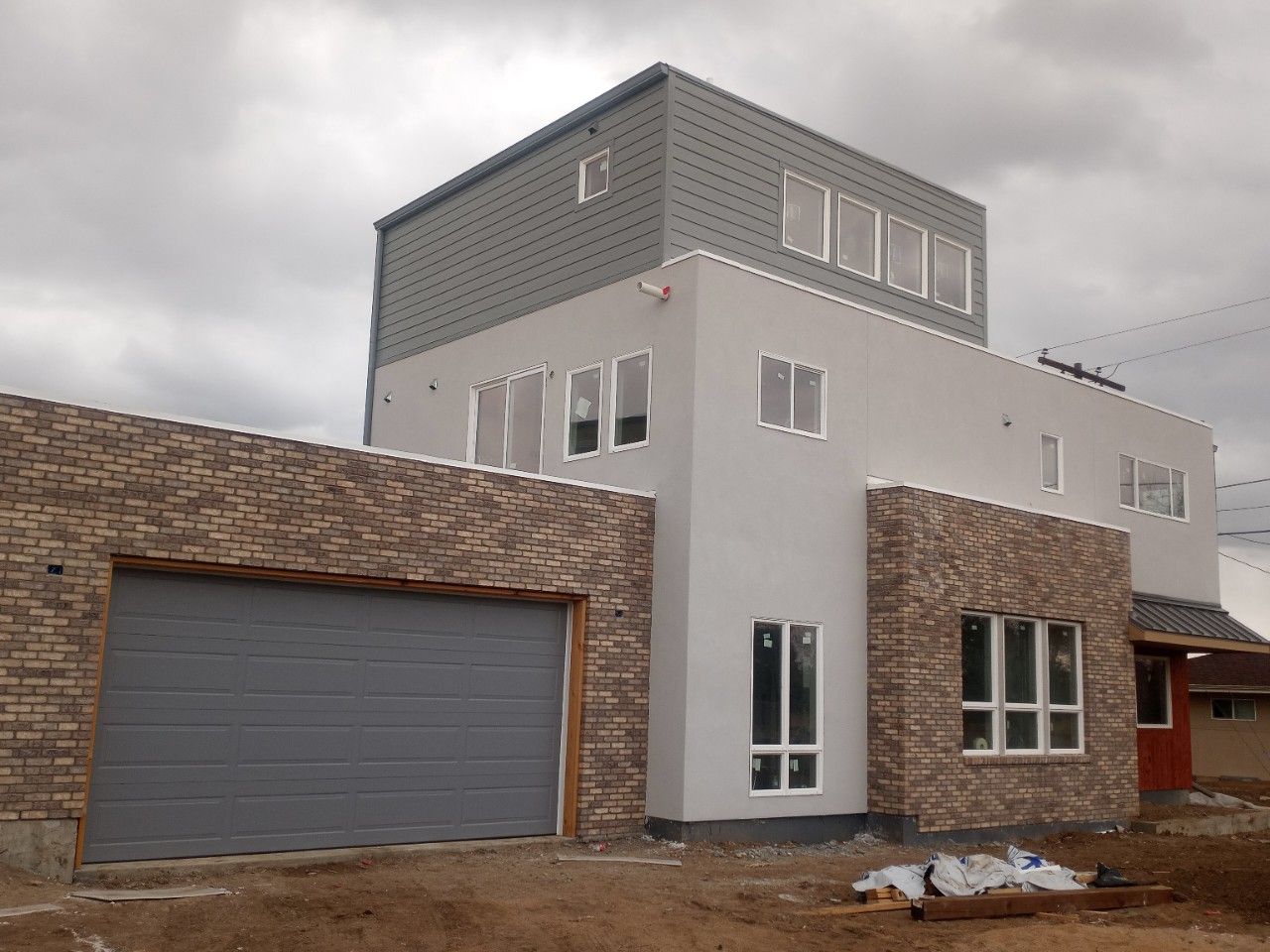 Modern two-story house under construction; gray garage door, brick accents, gray and white walls, and flat roof on cloudy day.