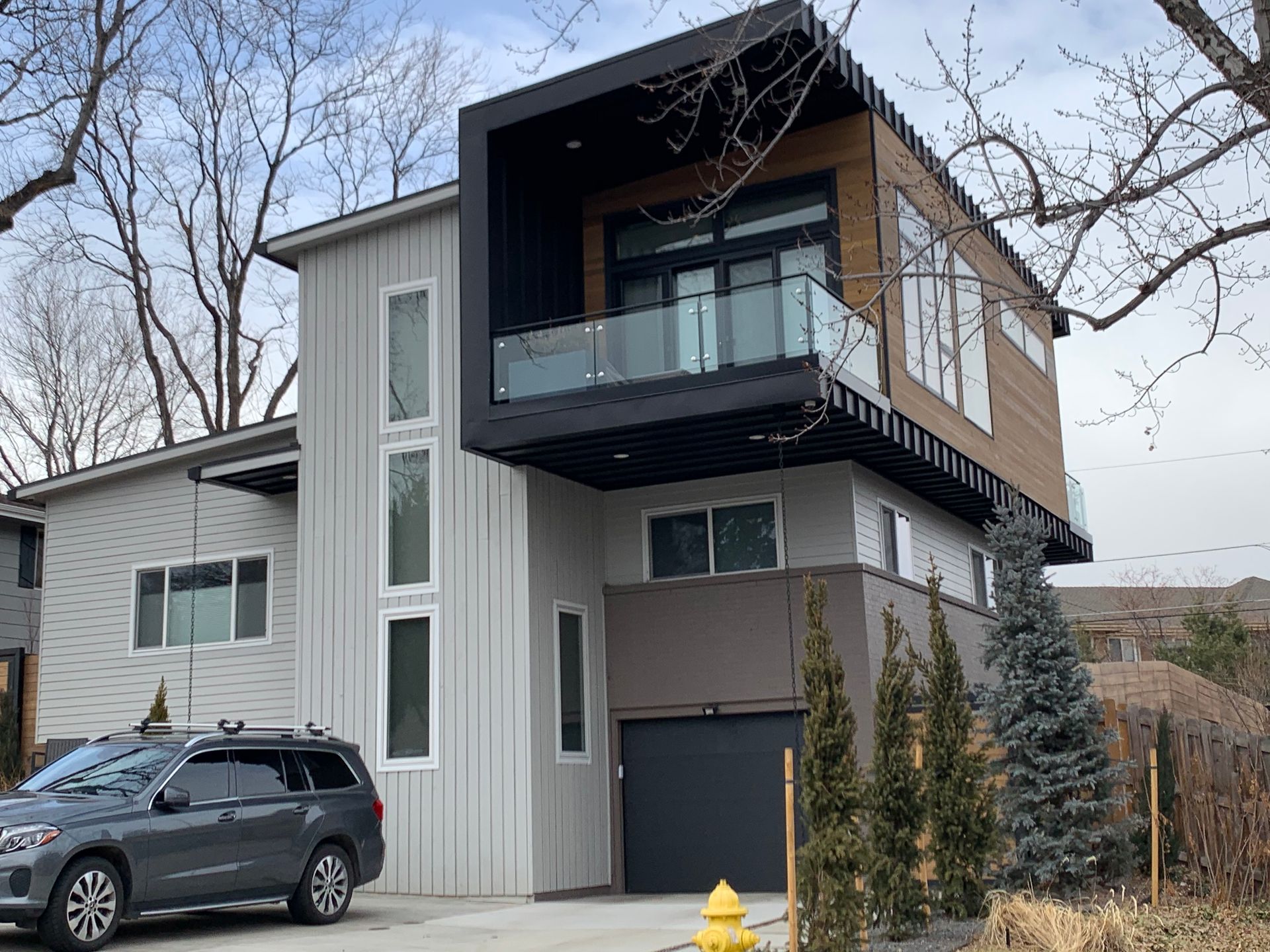 Modern two-story house with a protruding upper level featuring a balcony. Grey car parked in front.