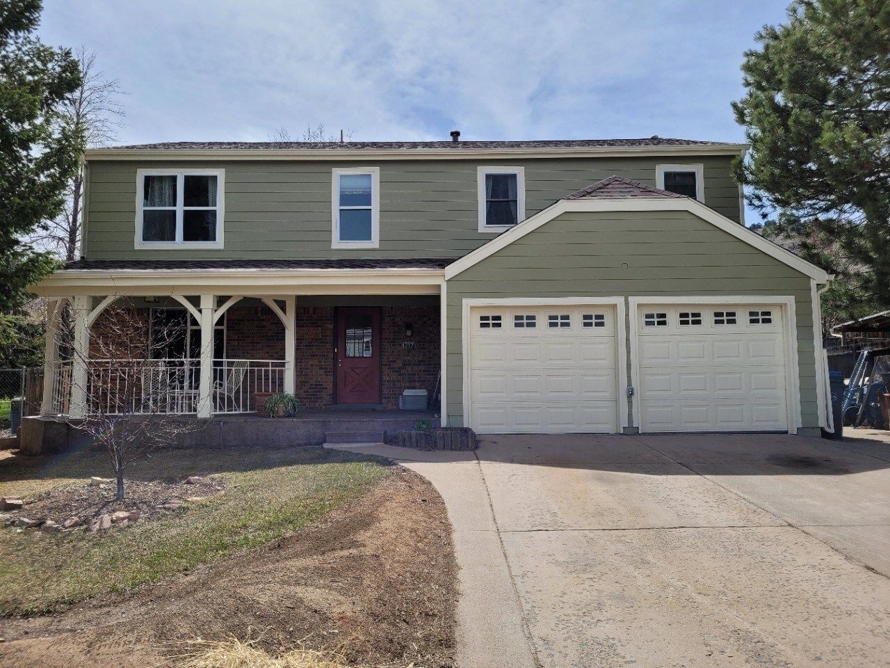 Two-story house with green siding, white garage doors, and a covered porch. A driveway leads to the house.