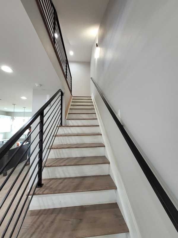 Staircase with light-colored wood steps and black metal railings ascending towards a hallway with a white wall.