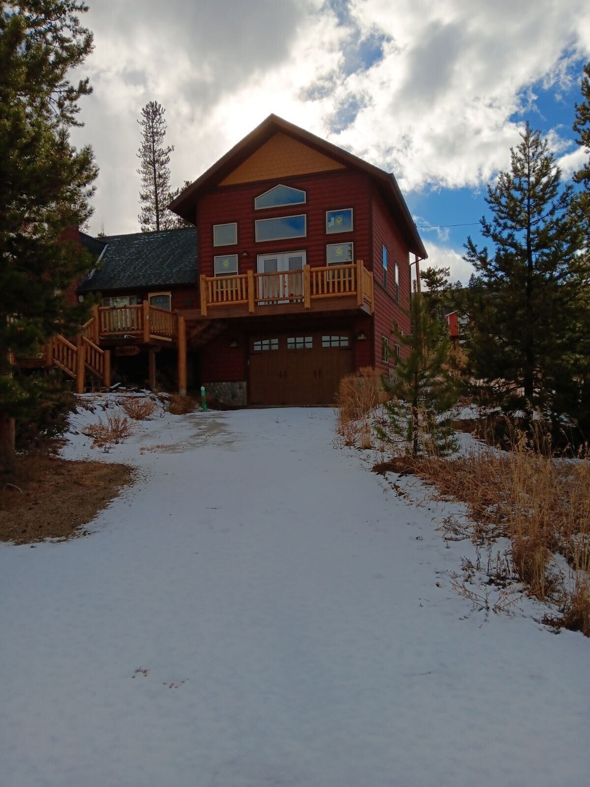 Red house with brown garage and deck, on a snow-covered driveway, surrounded by trees.
