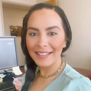 Woman with dark hair, smiling, wearing gold necklace, in an office setting.