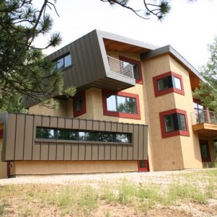 Modern multi-story home with tan stucco, angled brown metal siding, red window trim, and small balconies.