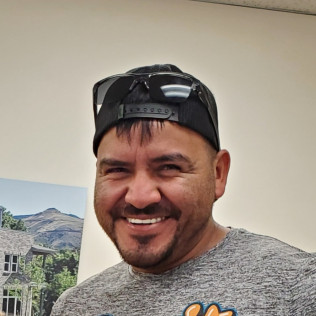 Man in cap and sunglasses smiles, wearing a gray shirt, indoors.