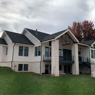 White house with black roof, brick accents, and green lawn under cloudy sky.