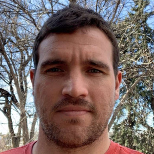 Man with short brown hair, beard, and red shirt, outdoors under a blue sky with leafless trees.