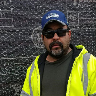 Man in sunglasses, hat, and yellow vest stands in front of a wire mesh background.