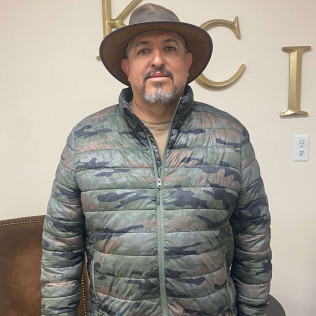 Man wearing a brown hat and camo jacket, standing in front of gold lettering.