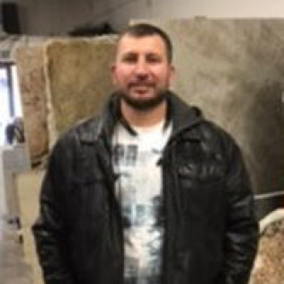 Man in a black leather jacket smiles, standing in front of stone slabs in a showroom.