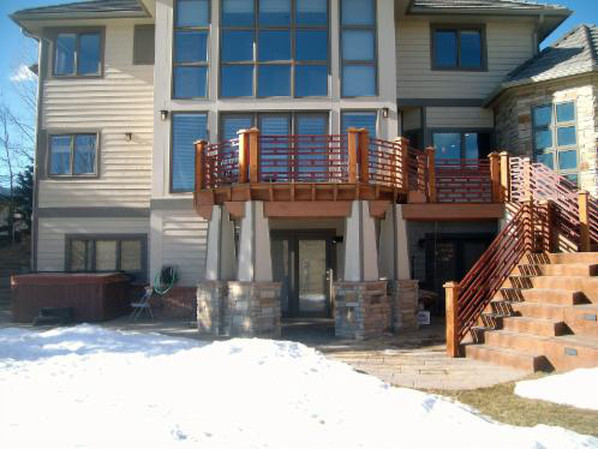 Two-story beige house with deck and stairs, supported by stone columns. Snow covers the ground.