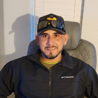 Man wearing a cap and jacket, seated in a chair, smiling at the camera.