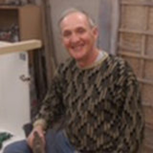 Smiling older man in patterned sweater, sitting in workshop.