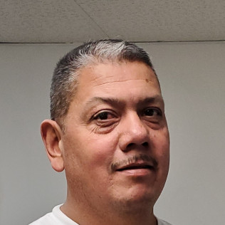 Man with short graying hair and mustache, looking at the camera, wearing a white shirt, in a room with a white wall.