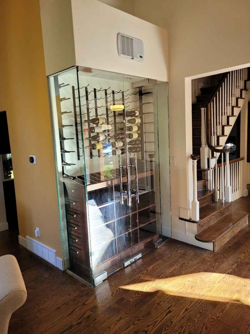 Glass-enclosed wine cellar with dark wood accents, shelves of bottles, and stairs in the background.
