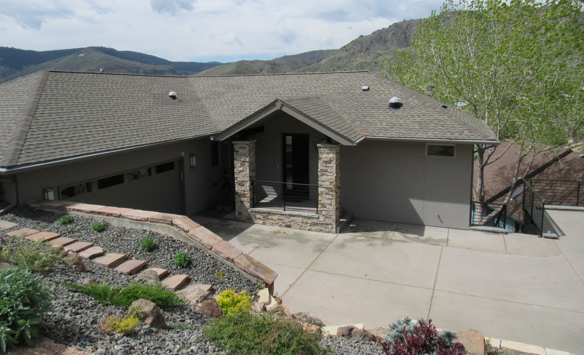 House with gray exterior, stone accents, and a sloped roof, driveway, and mountain backdrop.