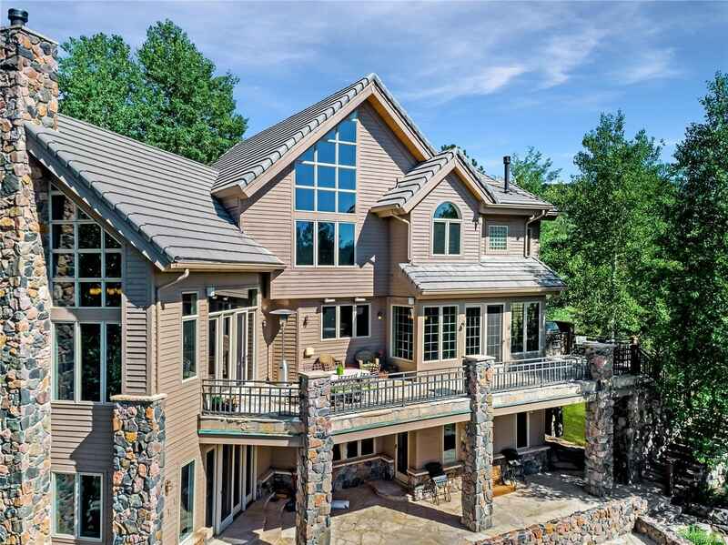 Large luxury house with stone accents and multiple balconies, surrounded by trees, under a blue sky.