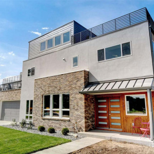 Modern, multi-story house with light-colored siding, brick accents, and a rooftop deck under a blue sky.
