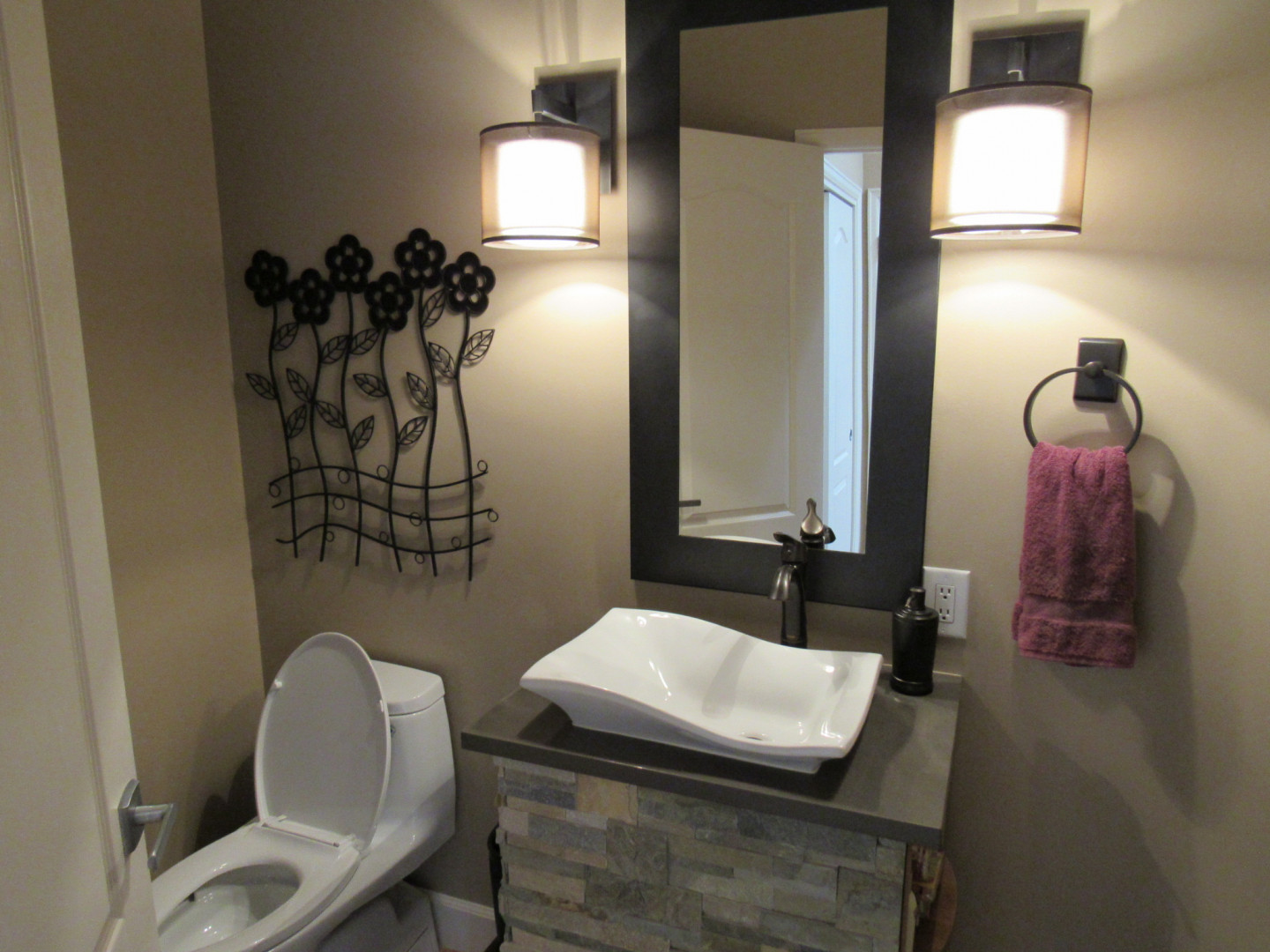 Bathroom with white sink, mirror, and toilet; tan walls, purple towel, and floral wall art.