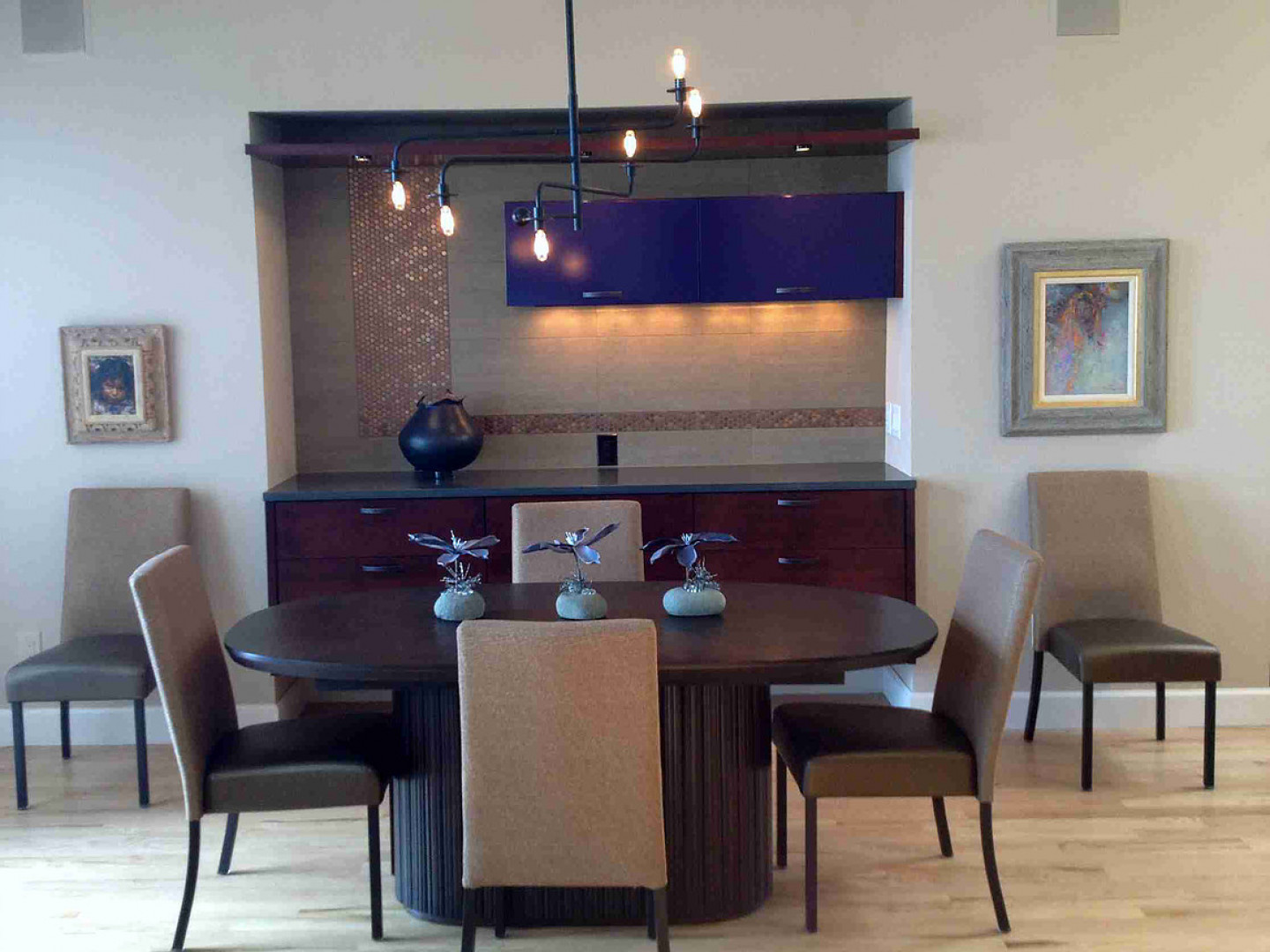 Dining room with oval table, chairs, built-in buffet, and artwork. Neutral tones and a dark chandelier.