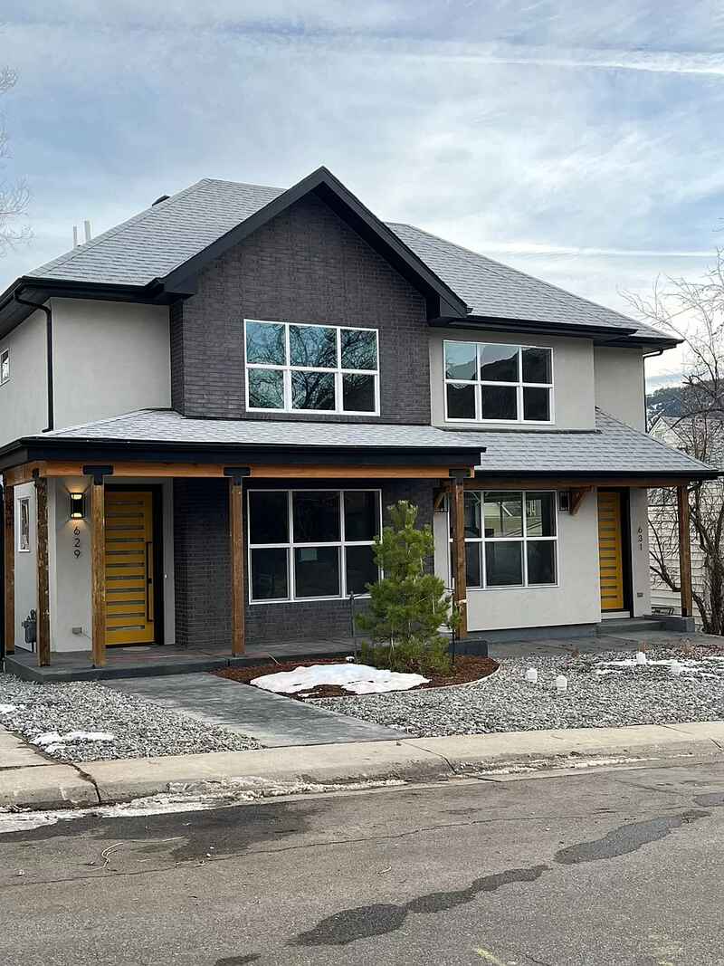 Two-story duplex with gray brick and light stucco exterior, yellow doors, and a small evergreen tree.