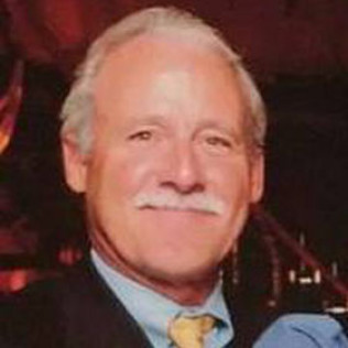 Man with white hair and mustache smiling in a dark setting, wearing a suit.