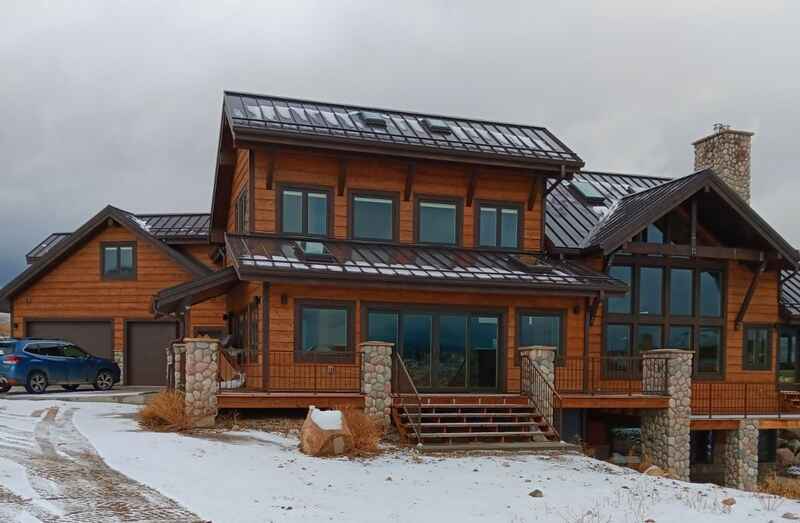 Rustic two-story log cabin home with a snowy driveway, blue car, and cloudy sky.