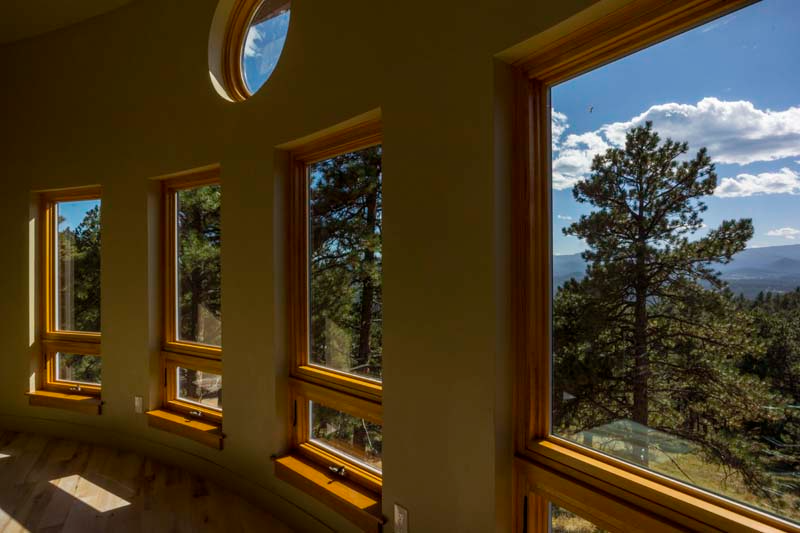 Interior view of curved wall with wooden-framed windows overlooking forest and mountains, blue sky.