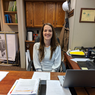Woman with long hair sits at a desk in an office, smiling. Papers and laptop in front of her.