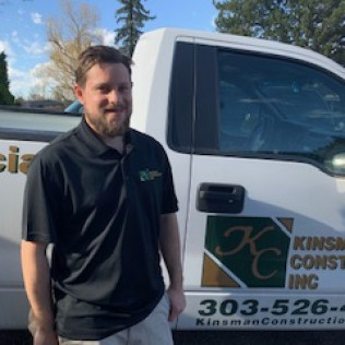 Man in black polo shirt, smiling, stands next to white Kinsman Construction truck with logo.