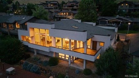 Modern two-story house with large windows and balcony, illuminated at dusk, in a suburban setting.