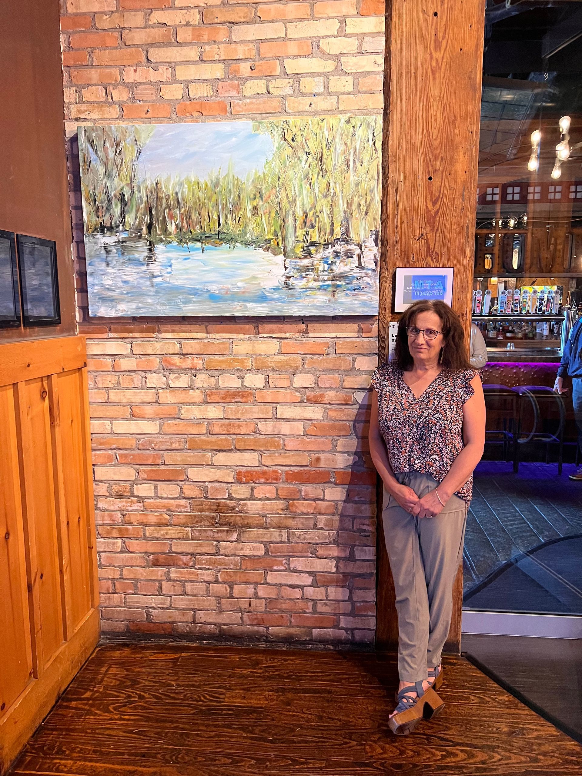 Woman stands by a brick wall with an abstract painting. She’s in a restaurant with a wooden post and bar in the background.
