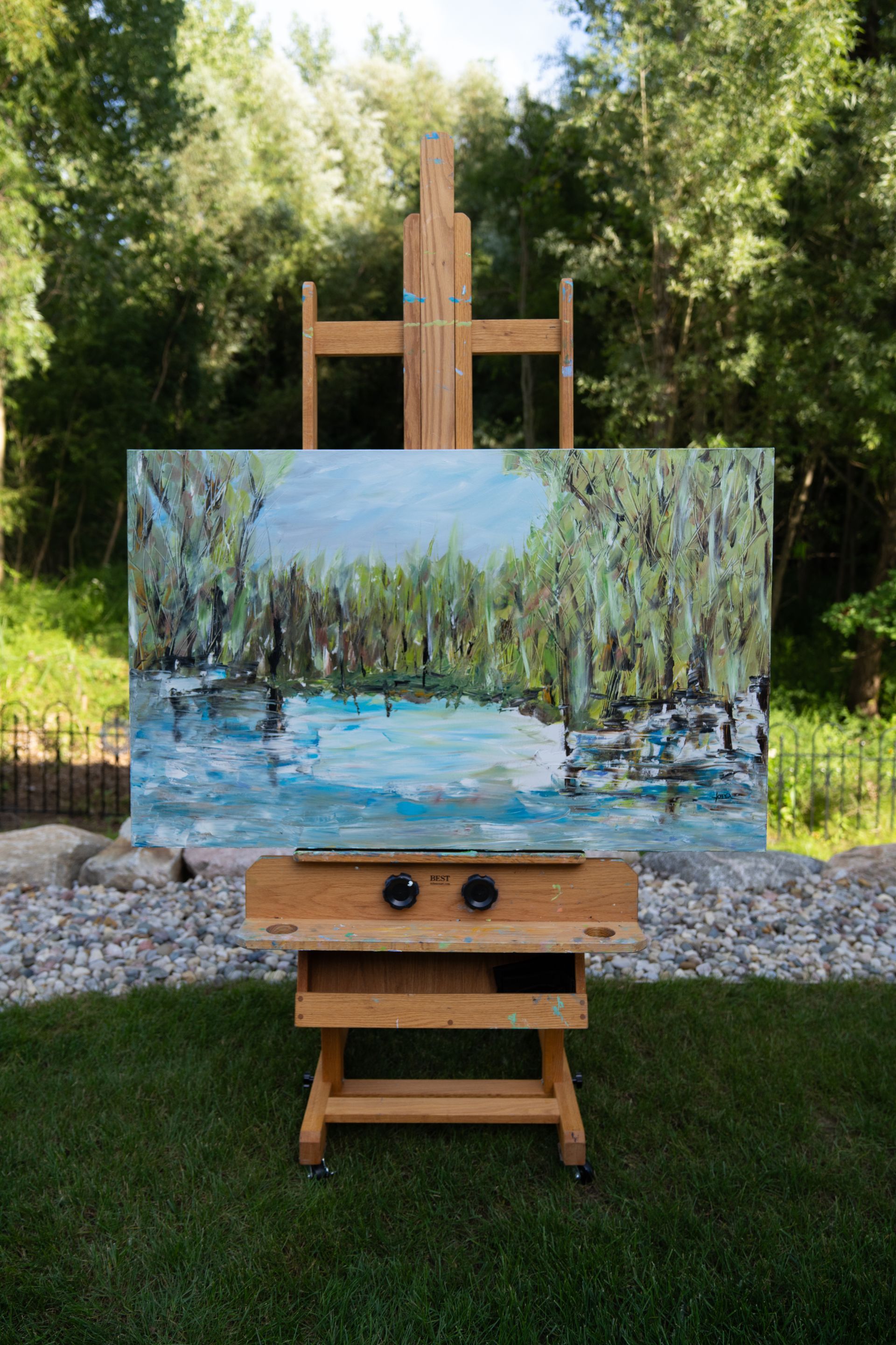 An abstract landscape painting on an easel outdoors, depicting a river scene in blues and greens.