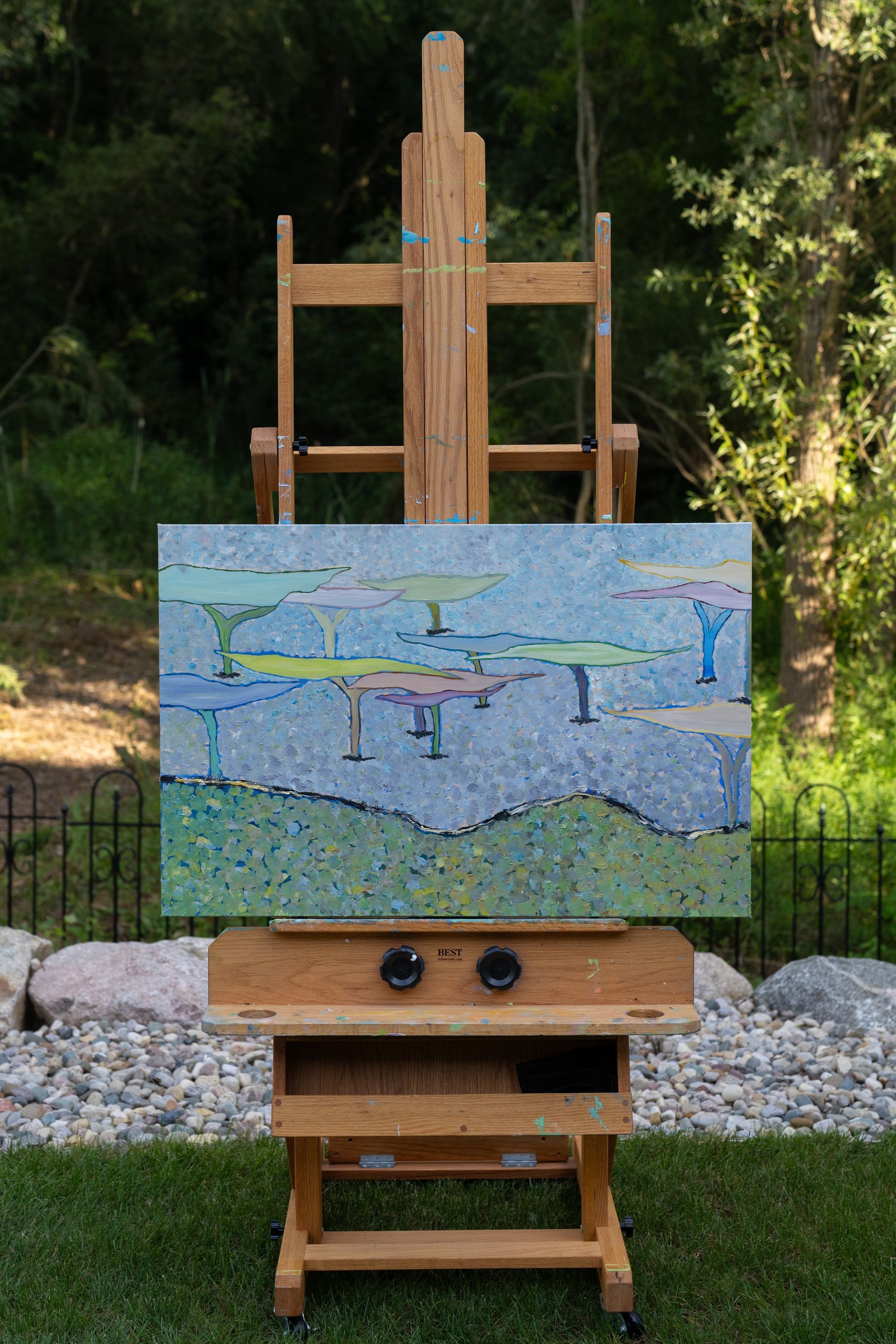 Painting on an easel outdoors; abstract landscape in blue, green, and yellow with a natural backdrop.